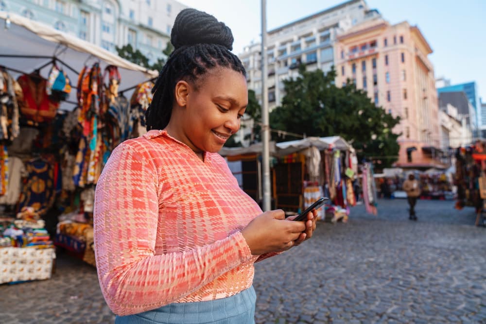 Woman in a pink top smiles while looking at her phone in an outdoor market with colorful stalls and buildings in the background.